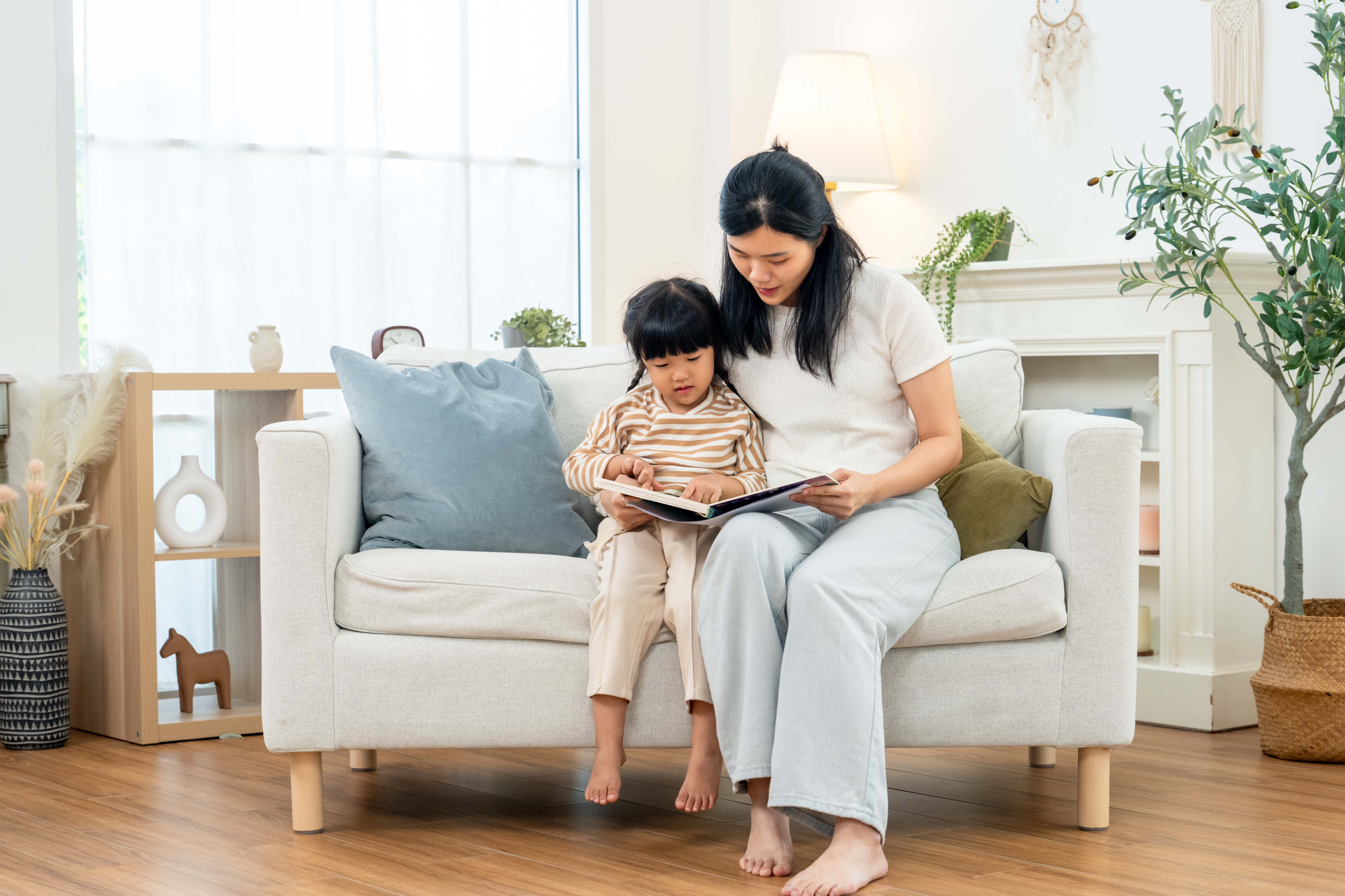 Asian mother sits with preschool daughter on sofa, turning picture book pages together in bright living room, warm family reading routine supporting bonding, language development, care, learning, play